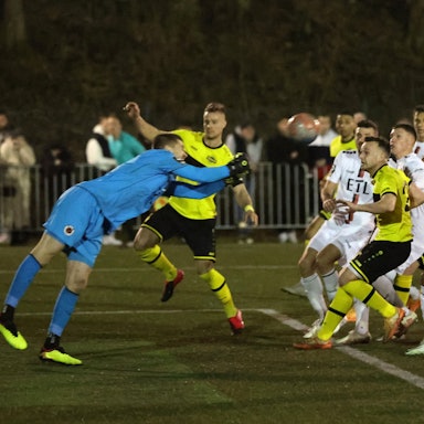 Ein Torwart in blauem Trikot faustet einen Ball zurück. Spieler in gelb-schwarzen Trikots und weißen Trickots nehmen ihn im Spiel wieder an.