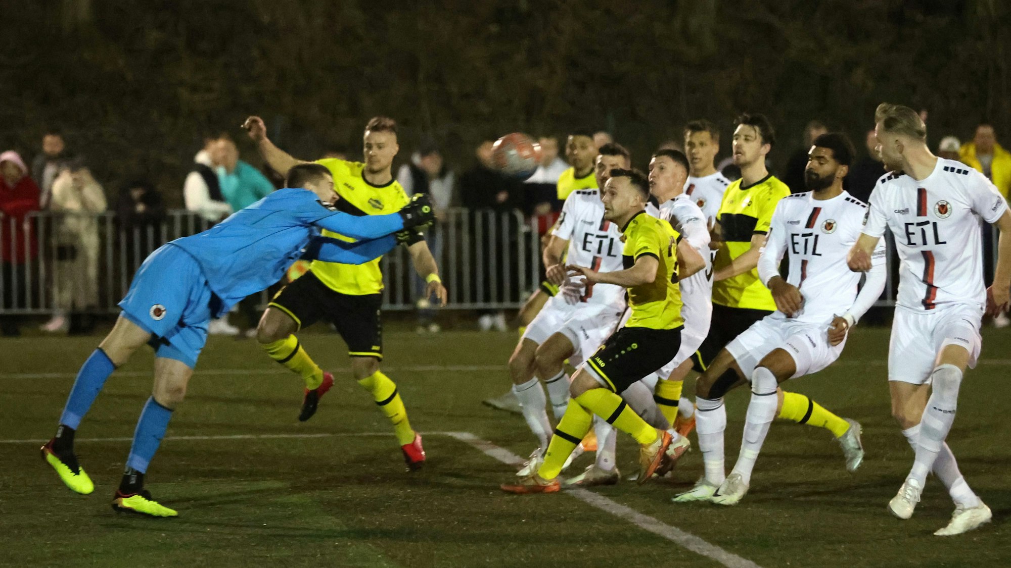 Ein Torwart in blauem Trikot faustet einen Ball zurück. Spieler in gelb-schwarzen Trikots und weißen Trickots nehmen ihn im Spiel wieder an.