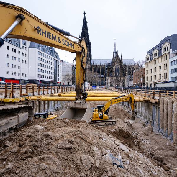 Zwei Baustellenfahrzeuge stehen in einem großen Loch und heben Erde aus. Im Hintergrund ist der Dom zu sehen.