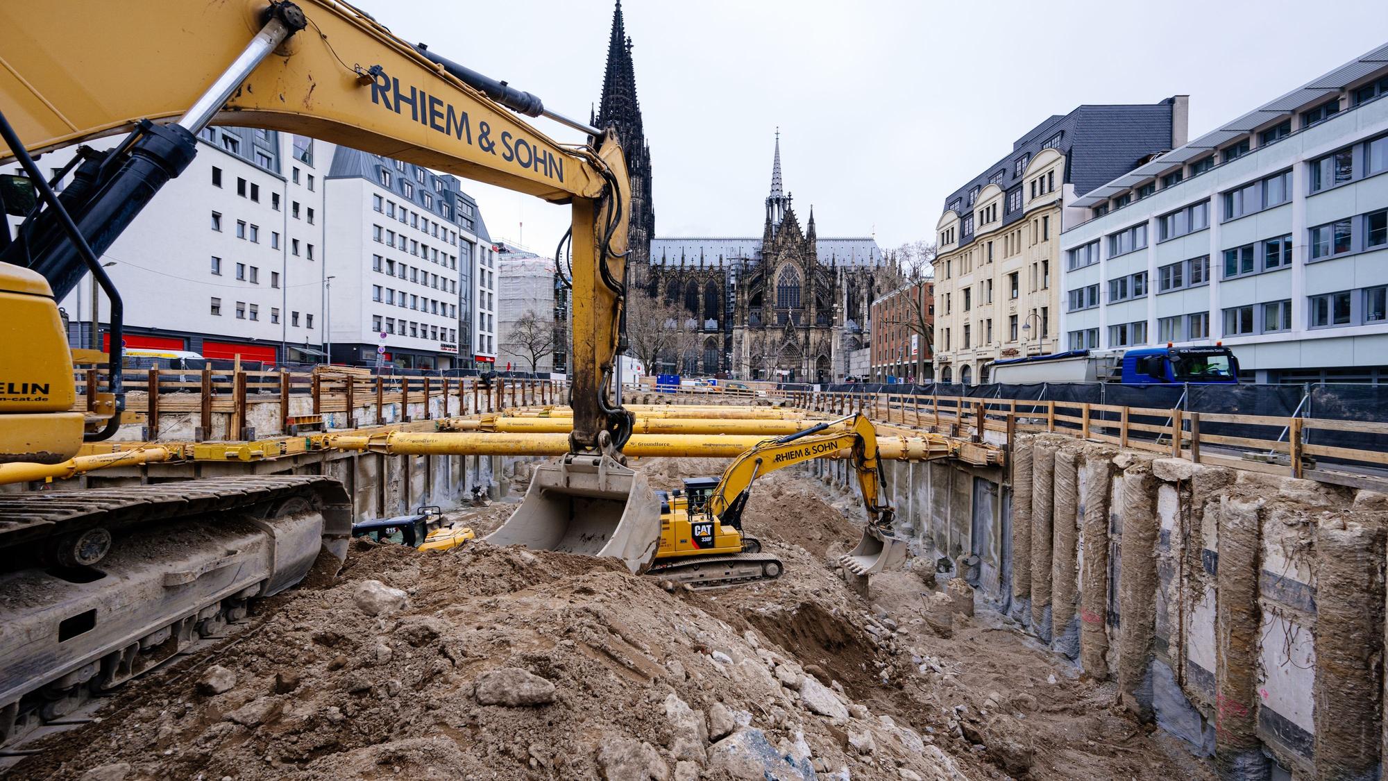 Zwei Baustellenfahrzeuge stehen in einem großen Loch und heben Erde aus. Im Hintergrund ist der Dom zu sehen.