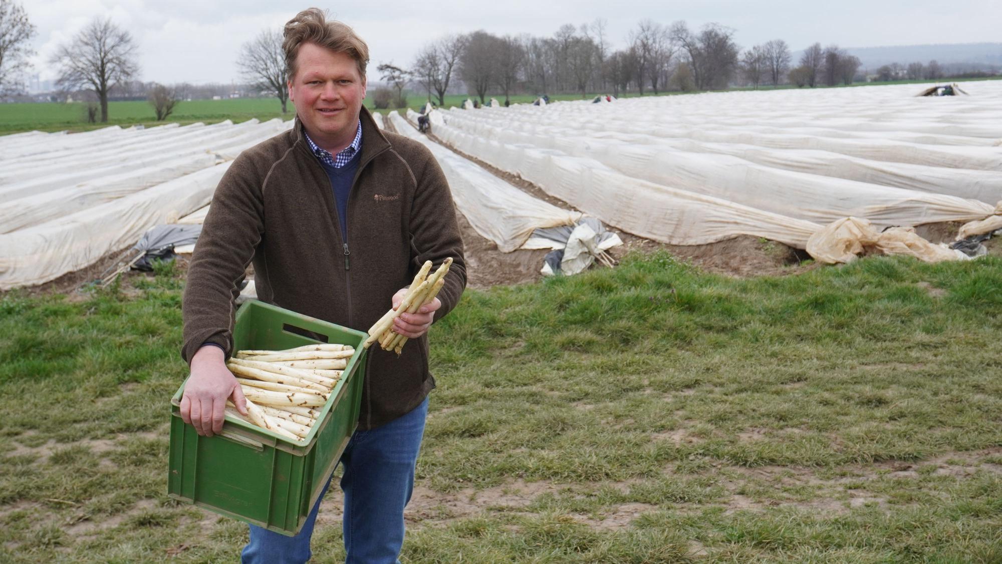 Auf dem Foto ist Johannes Nagelschmitz zu sehen. Er steht vor einem Spargelfeld und trägt eine grüne Kiste mit frisch geerntetem Spargel.