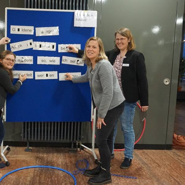 Andreas Ickes, Jana Wiens, Heike Heinen und Svenja Pommer-Schmitt stehen vor einer blauen Tafel, welche das Konzept für den "Medienparcours" für dritte und vierte Klassen präsentiert.