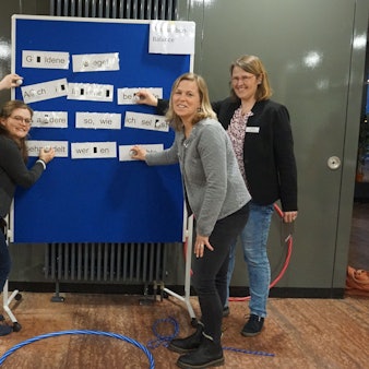Andreas Ickes, Jana Wiens, Heike Heinen und Svenja Pommer-Schmitt stehen vor einer blauen Tafel, welche das Konzept für den "Medienparcours" für dritte und vierte Klassen präsentiert.