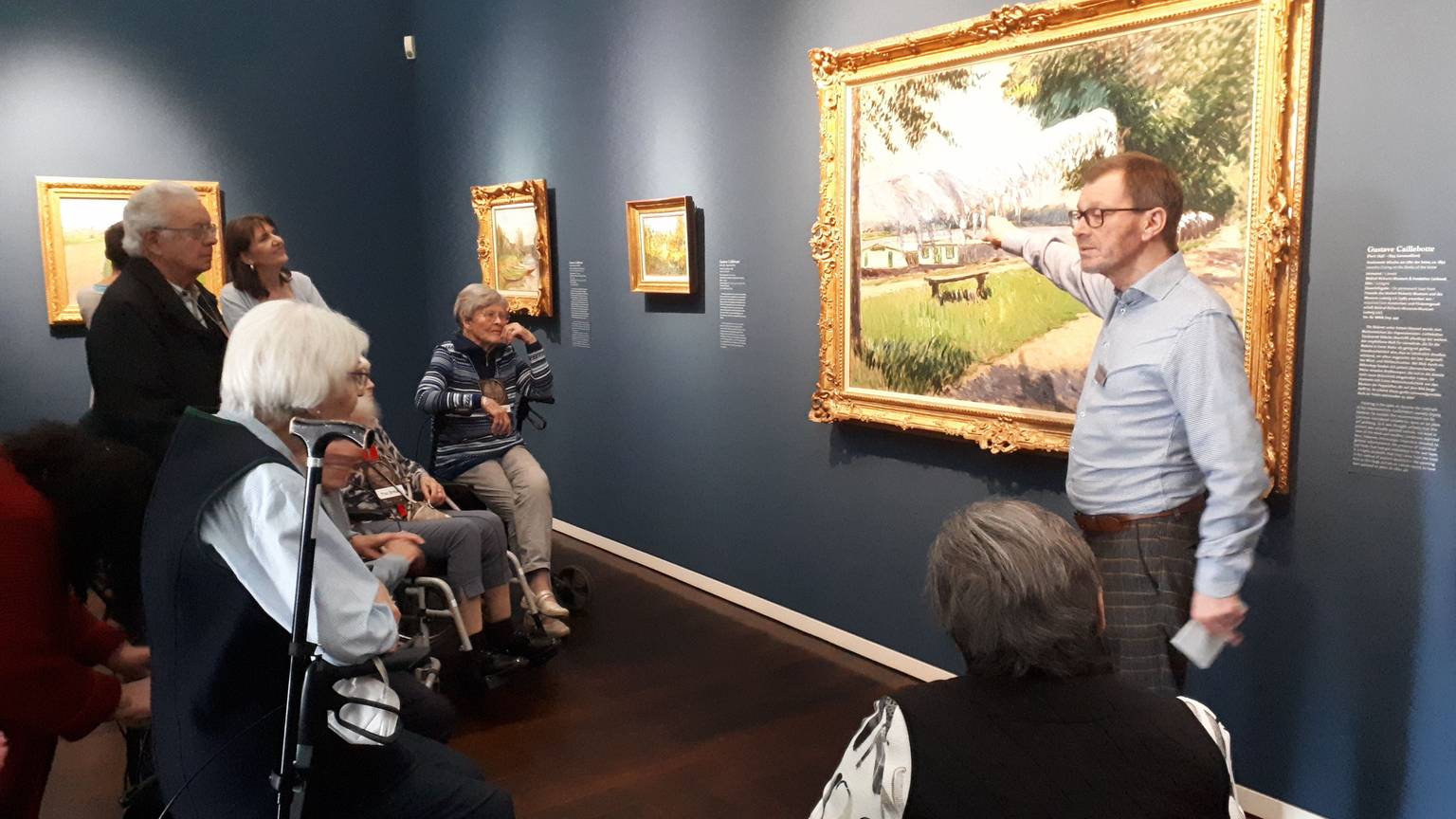 Ein Mann beschreibt ein Kunstwerk in einem Museum. Seniorinnen und Senioren schauen ihm zu.