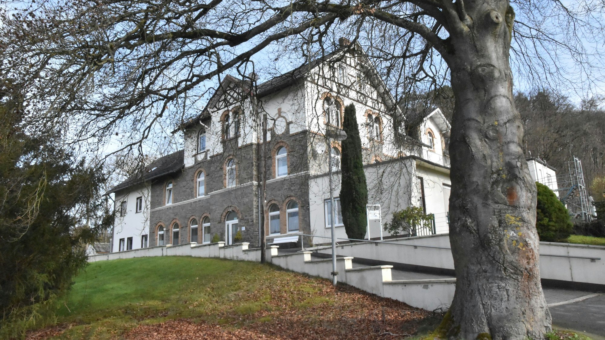 Unter einem großen Baum auf einem grünen Hügel ist das ehemalige Schullandheim und heutige Seniorenheim zu erkennen: Ein Gebäude aus Natursteinen, weißem Gemäuer und Fachwerk sowie einer breiten Zufahrt an der Seite.