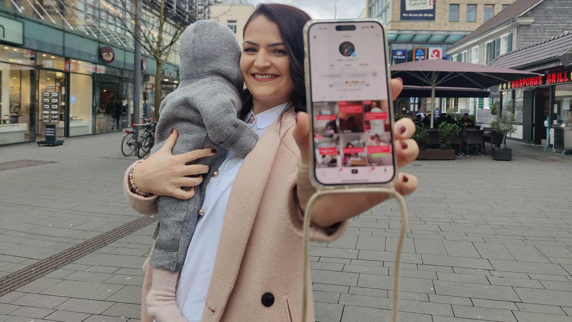 Die Bergisch Gladbacherin Mumfluencerin hält ihr Smartphone in die Kamera, ihr Kind träge sie auf dem anderen Arm.