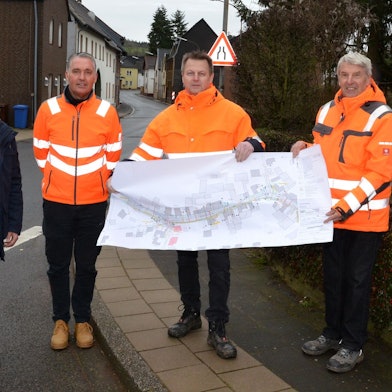 Drei Männer mit orangen Warnwesten halten einen Stadtplan hoch. Daneben steht ein Mann in Straßenklamotten.