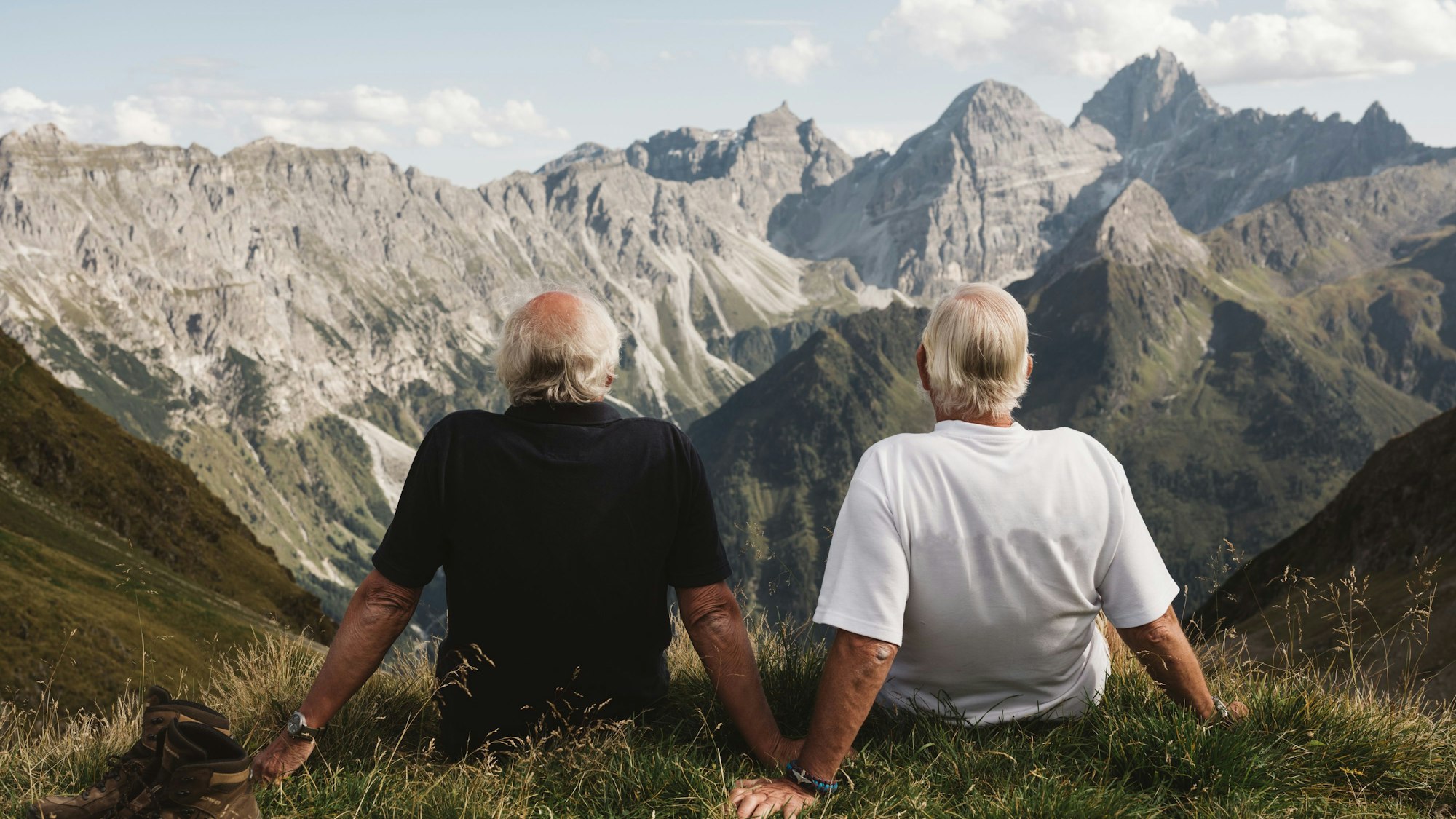 Zwei ältere Menschen sitzen an einem Hang und schauen auf eine Berglandschaft hinaus.
