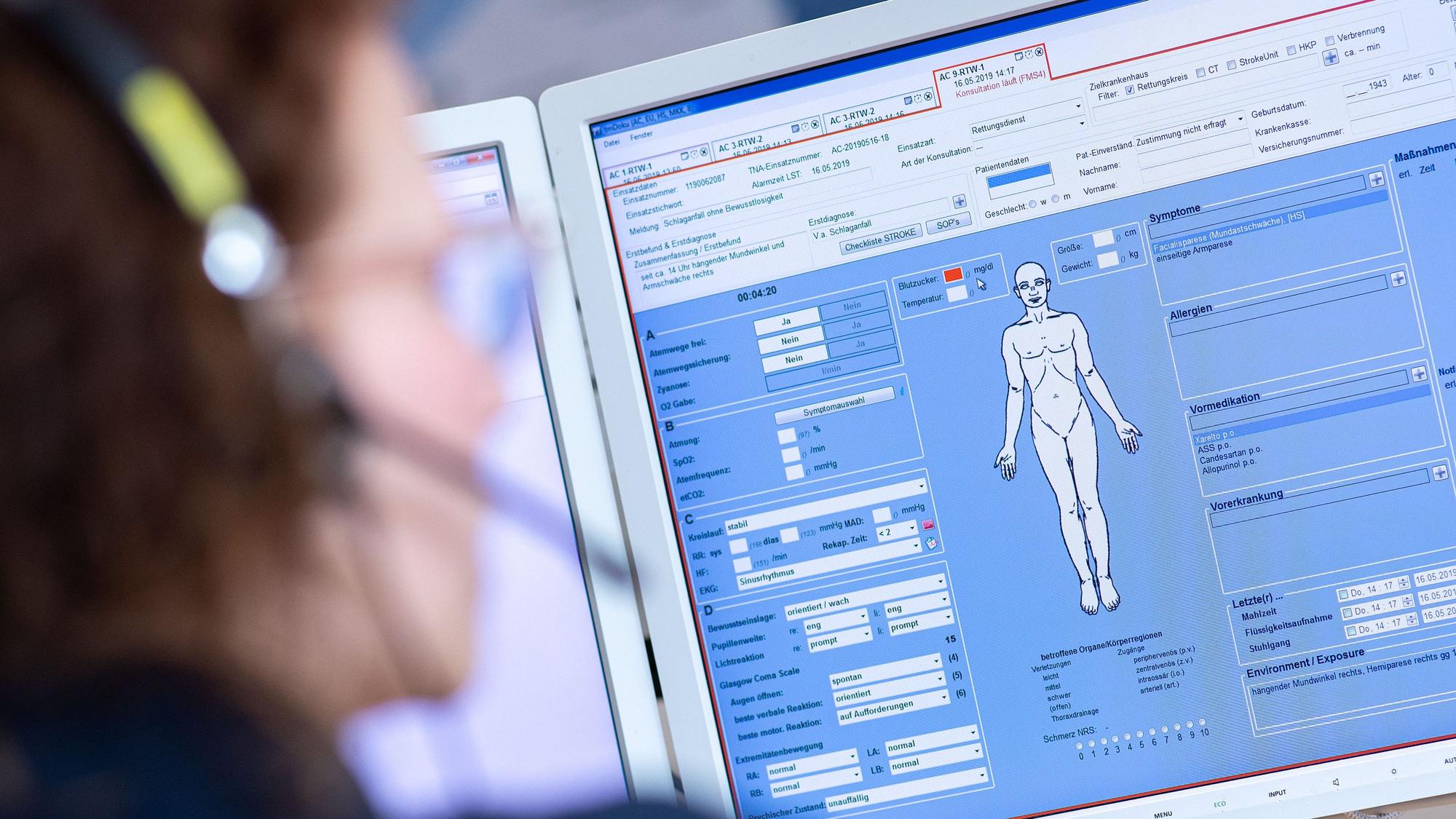 Eine Telenotärztin sitzt in der Rettungsleitstelle vor einem Bildschirm und ist mit Rettungssanitätern verbunden, die eine Schlaganfallpatientin behandeln.