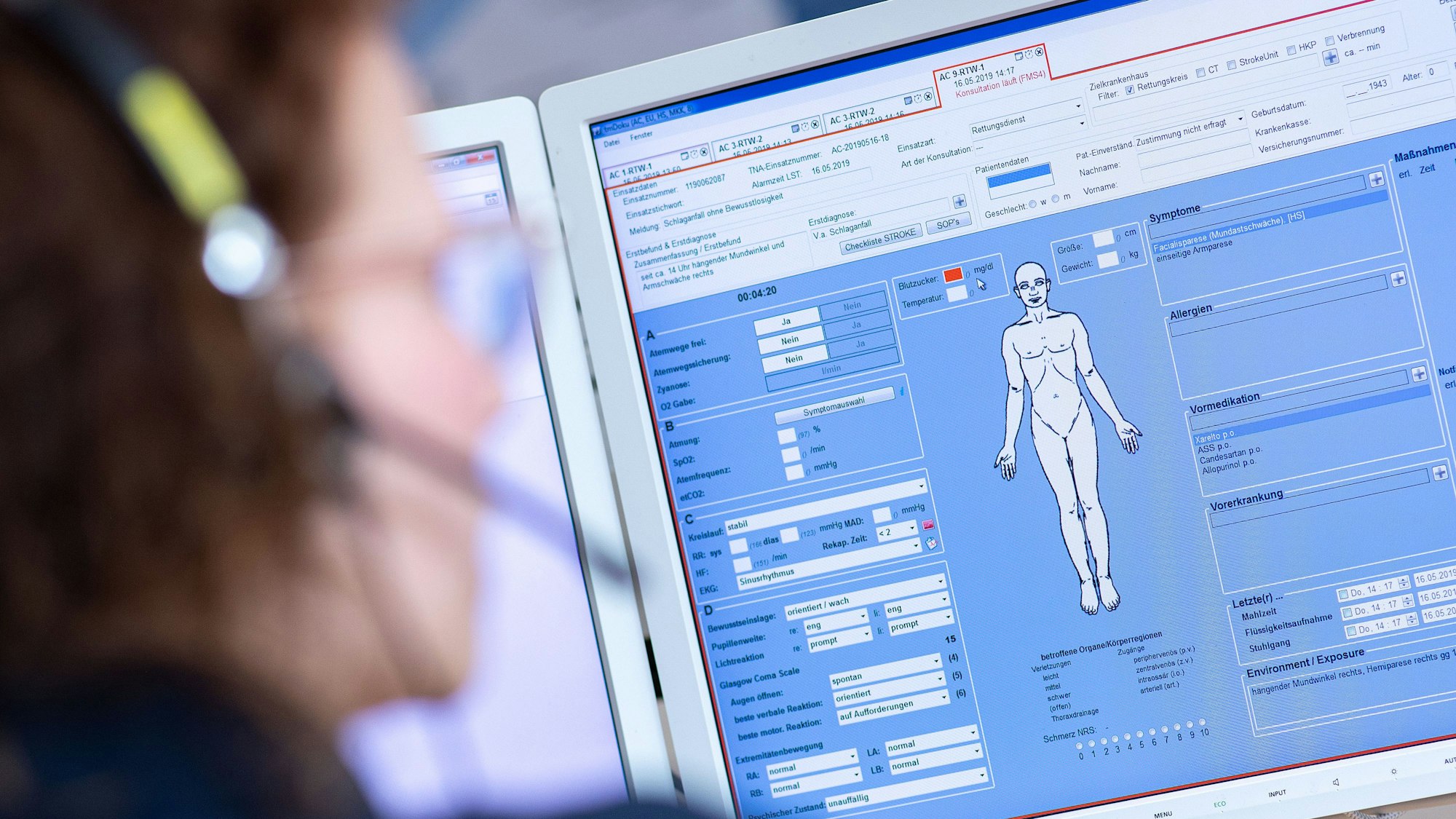 Eine Telenotärztin sitzt in der Rettungsleitstelle vor einem Bildschirm und ist mit Rettungssanitätern verbunden, die eine Schlaganfallpatientin behandeln.