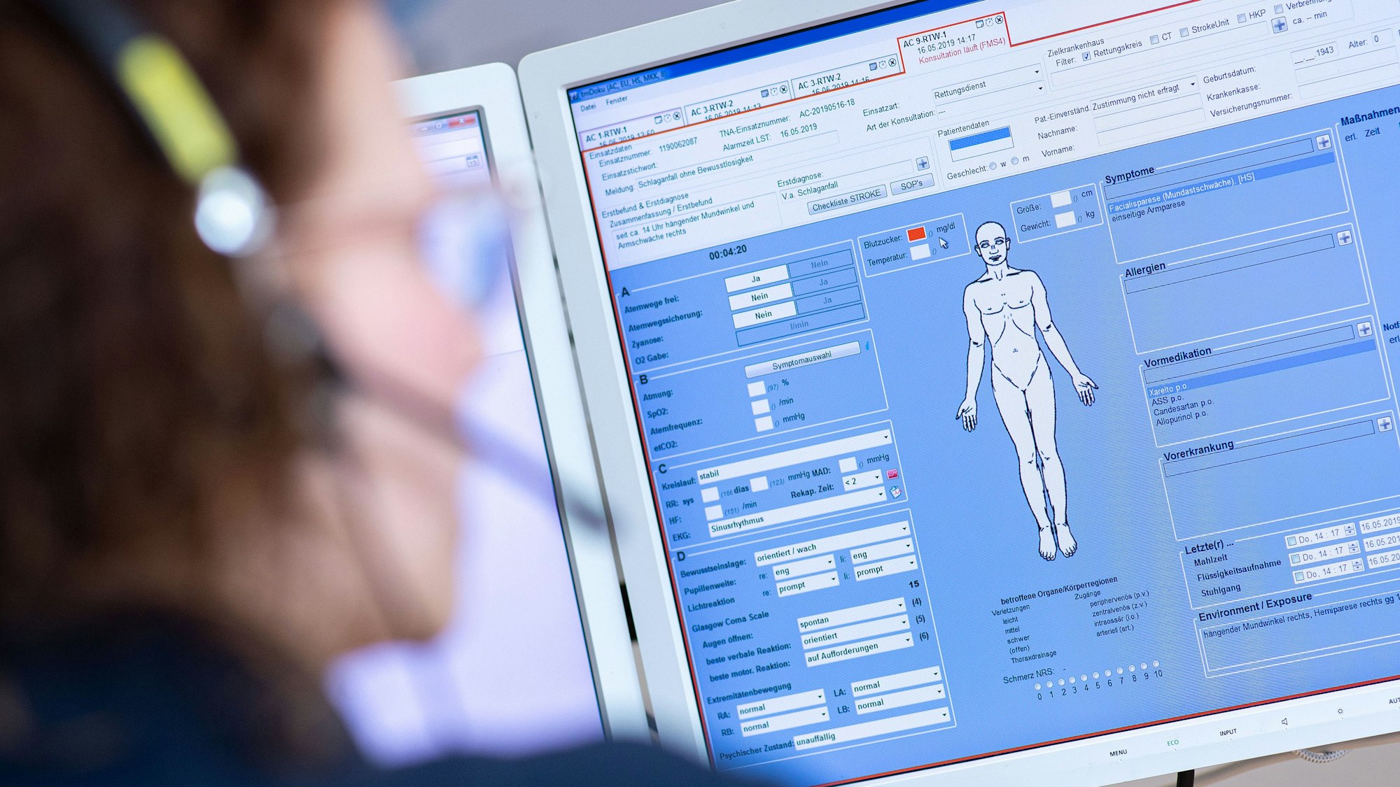 Eine Telenotärztin sitzt vor einem Bildschirm und ist mit Rettungssanitätern verbunden, die eine Schlaganfallpatientin behandeln.