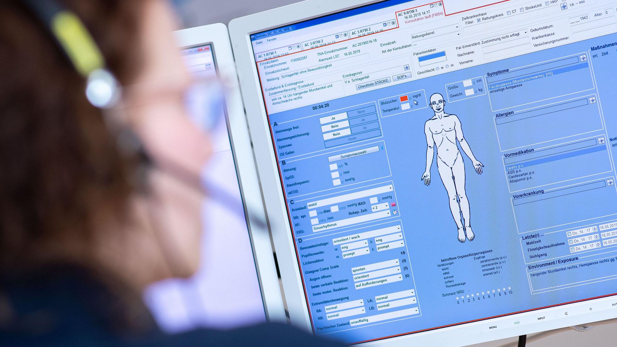 Eine Telenotärztin sitzt in der Rettungsleitstelle der Feuerwehr vor einem Bildschirm und ist mit Rettungssanitätern verbunden, die eine Schlaganfallpatientin behandeln.