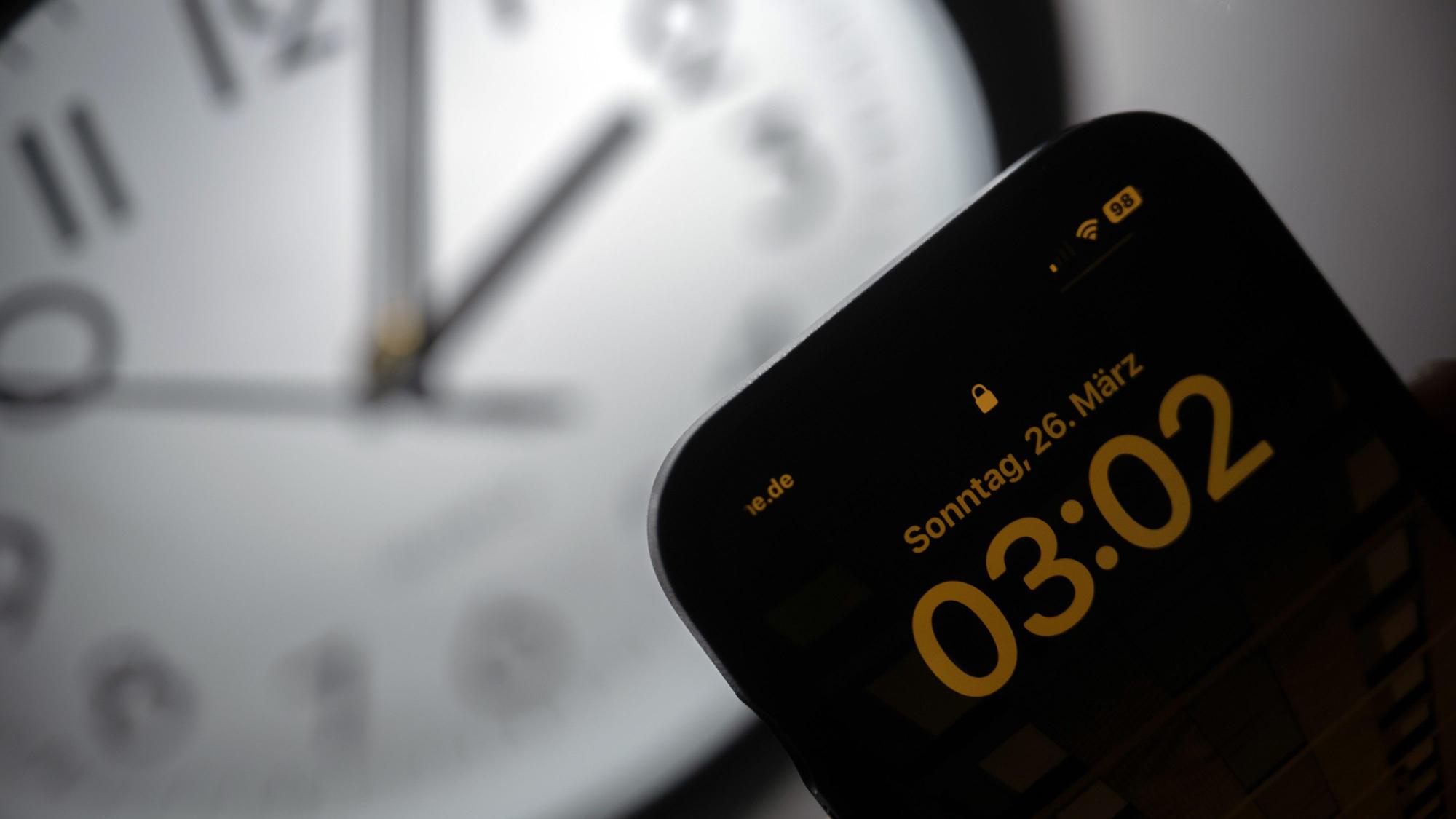 Ein Smartphone zeigt die Uhrzeit «03:03» an, während auf einer analogen Uhr noch 02:03 Uhr ist.