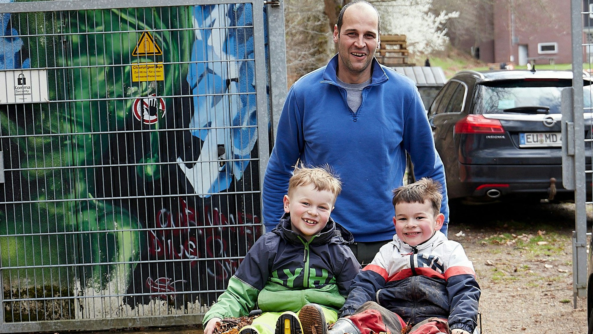Die beiden Kinder sitzen auf dem Laub in der Schubkarre und werden von ihrem Vater weggefahren.