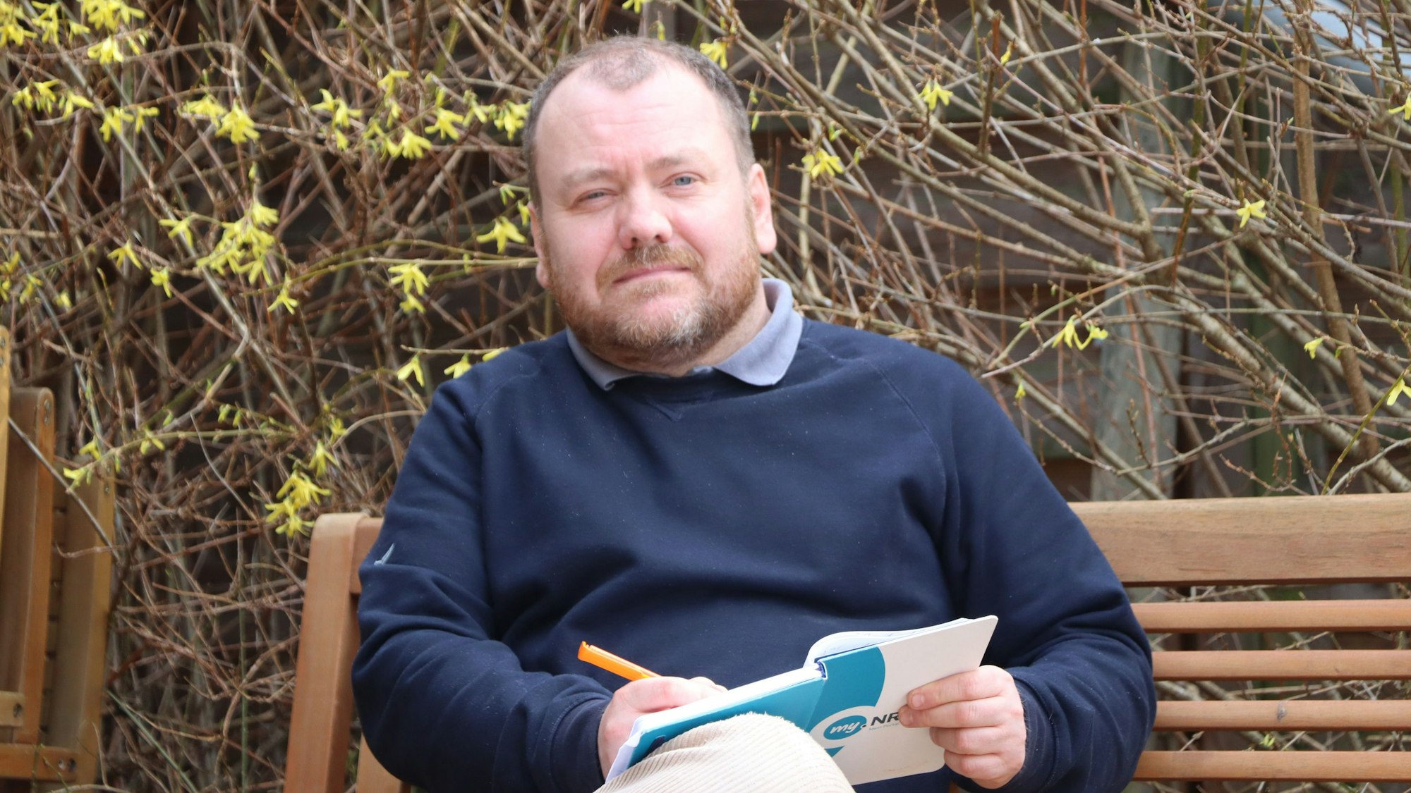 Ein Mann mit einem Notizbuch in der Hand sitzt auf einer Bank.