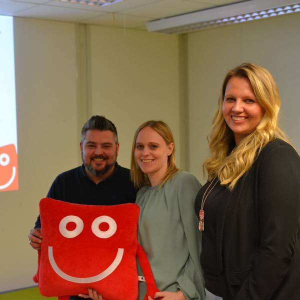 Die Leiter halten das rote, eckige Stoffmaskottchen in den Händen und lächeln in die Kamera.
