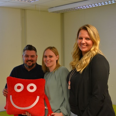Die Leiter halten das rote, eckige Stoffmaskottchen in den Händen und lächeln in die Kamera.