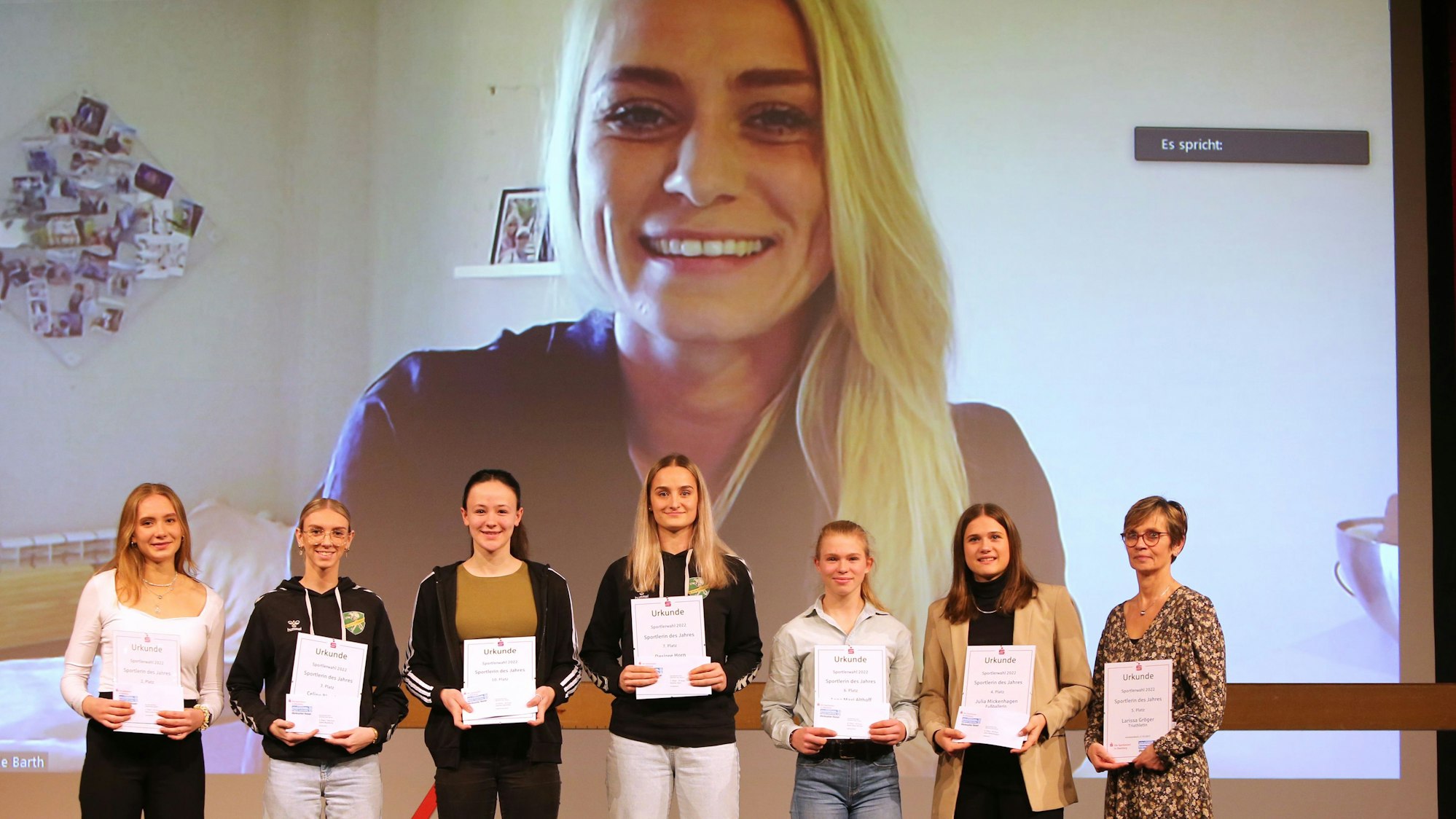 Blick auf eine Bühne, auf der Sportlerinnen stehen und über eine Leinwand gezeigt werden.