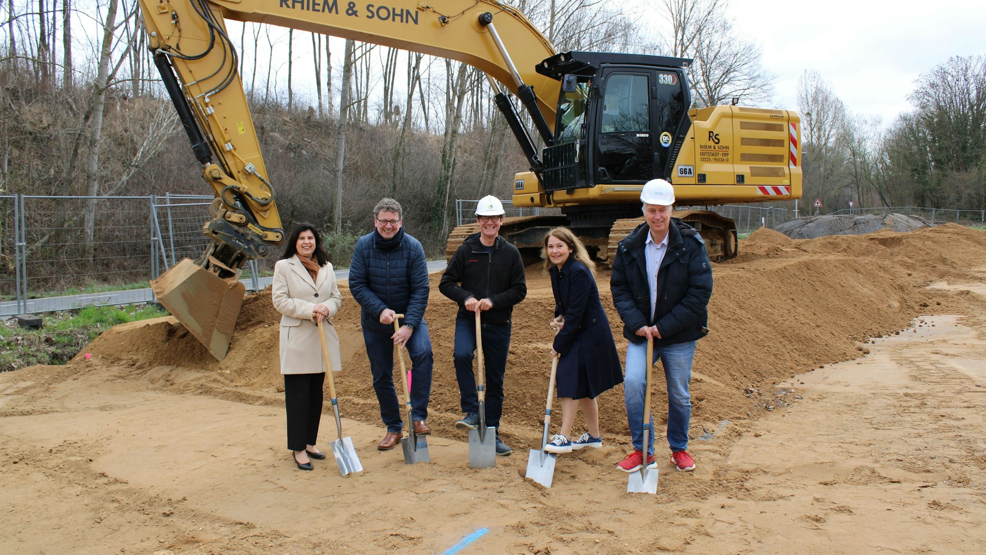 Die fünf Personen stehen vor einem Bagger und halten Spaten in der Hand.