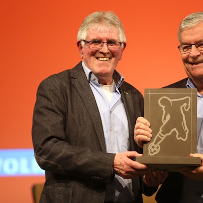 Zwei Männer stehen nebeneinander. In der Mitte halten sie gemeinsam einen eckigen Stein-Pokal in der Hand, in dessen Platte ein Fußballer eingraviert ist.