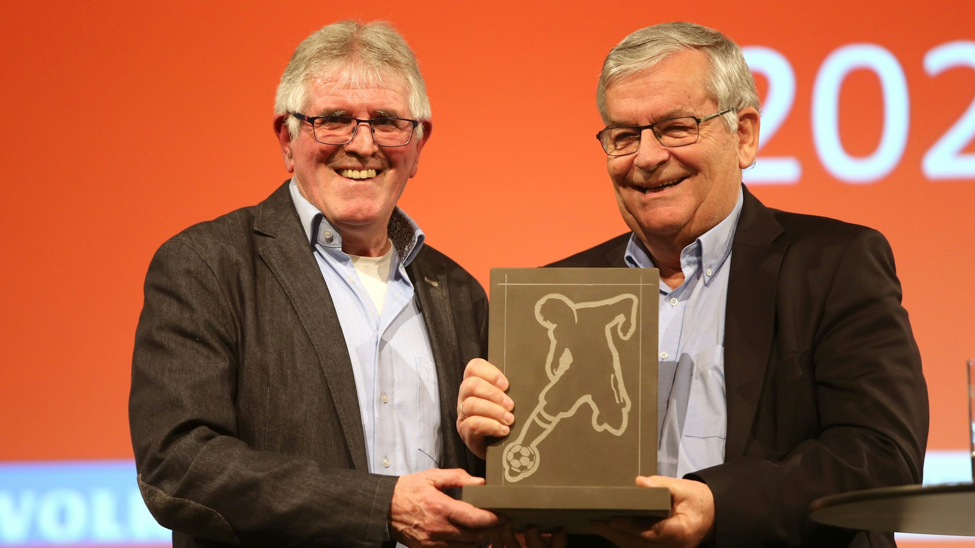 Zwei Männer stehen nebeneinander. In der Mitte halten sie gemeinsam einen eckigen Stein-Pokal in der Hand, in dessen Platte ein Fußballer eingraviert ist.