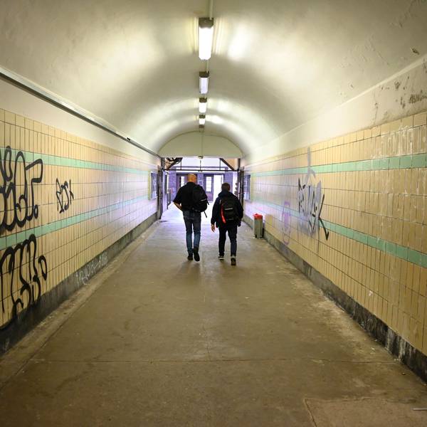 Zu sehen sind zwei Männer, die durch die Unterführung eines Bahnhofs gehen, deren Wände beschmiert sind.