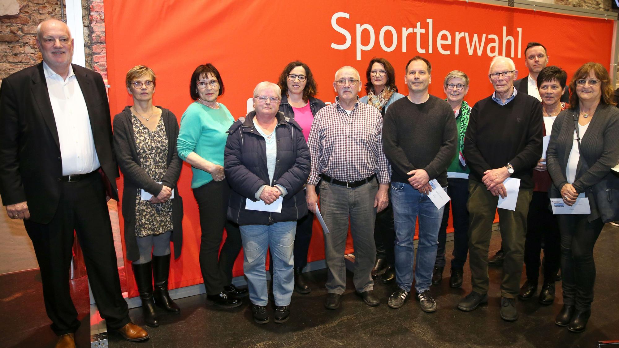 Auf einem Gruppenfoto stehen mehrere Frauen und Männer gemeinsam vor einem roten Banner mit der weißen Aufschrift „Sportlerwahl“.