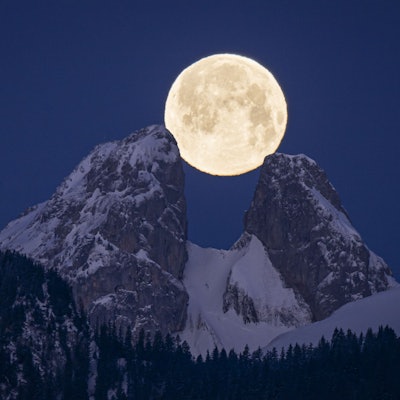 Der Vollmond geht hinter zwei verschneiten Berggipfeln auf. Er ist deutlich am klaren und tiefblauen Himmel zu sehen.