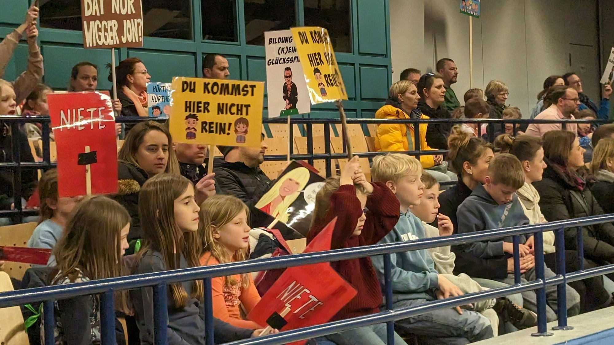 Eltern und Kinder sitzen im Publikum der Ratssitzung im Römersaal des Bürgerhauses. Einige halten Schilder mit Aufschriften wie „Niete“, „Du kommst hier nicht rein!“ oder „Wie soll dat nur wigger jon?“.