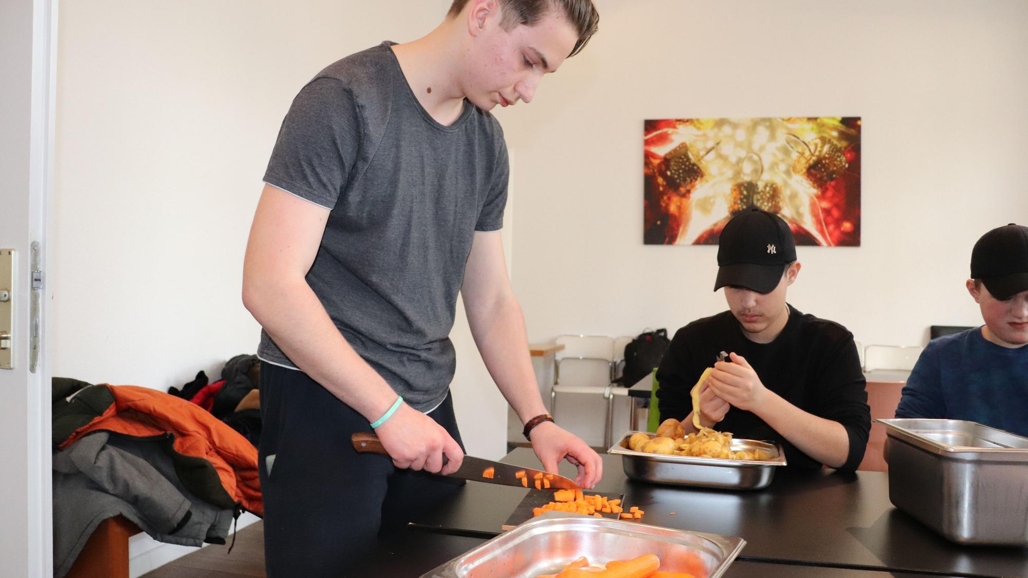 Zwei Jungen sind dabei zu sehen, eine Möhre zu schneiden und eine Kartoffel zu schälen.
