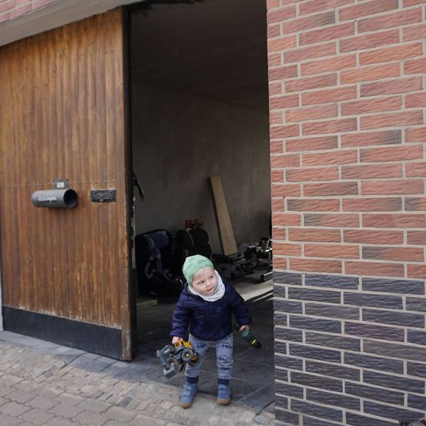 Der kleine Max, einen Spielzeugbagger in der Hand, lugt vorsichtig aus dem Hoftor auf die Straße vor dem Haus.