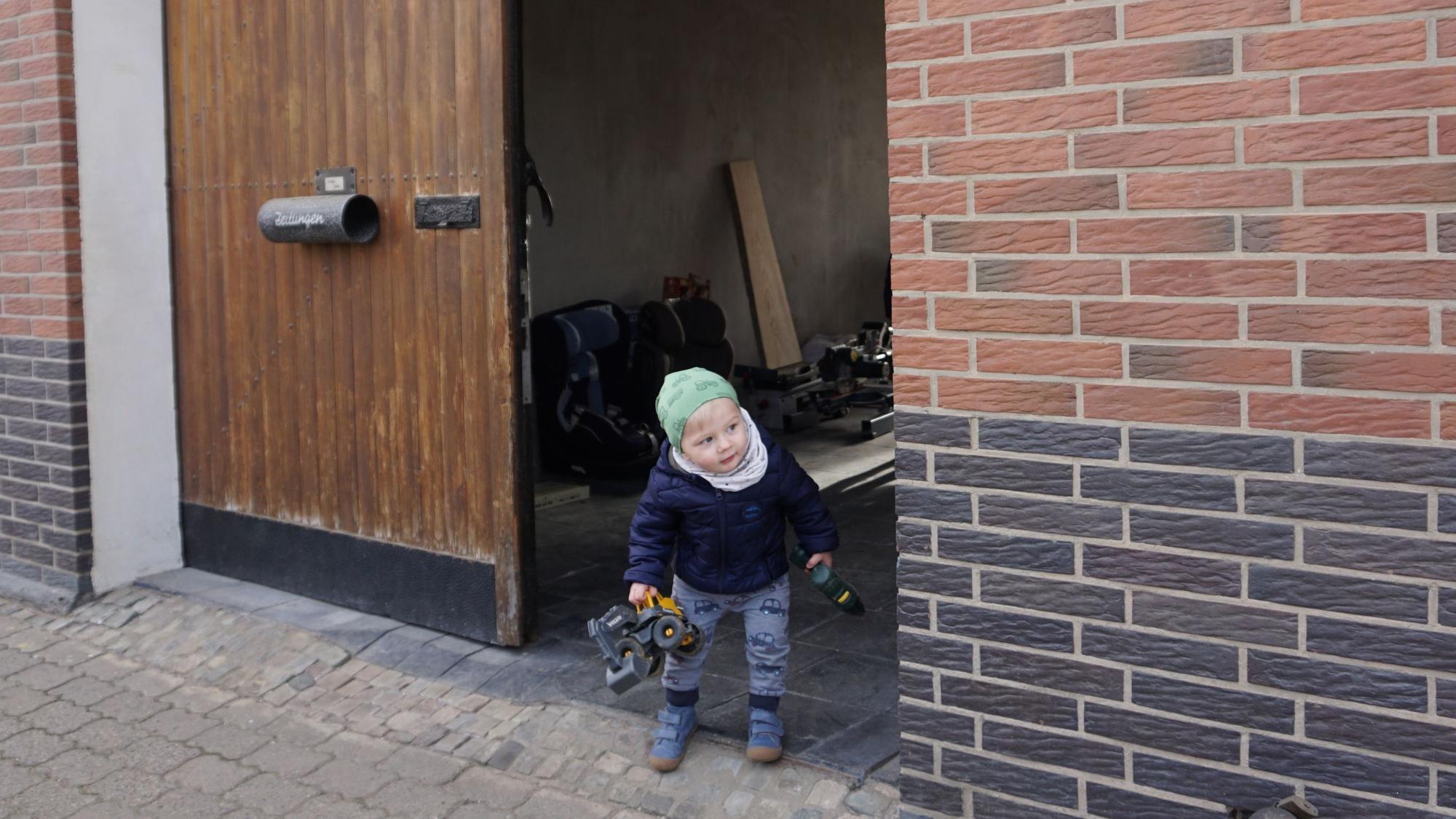 Der kleine Max, einen Spielzeugbagger in der Hand, lugt vorsichtig aus dem Hoftor auf die Straße vor dem Haus.