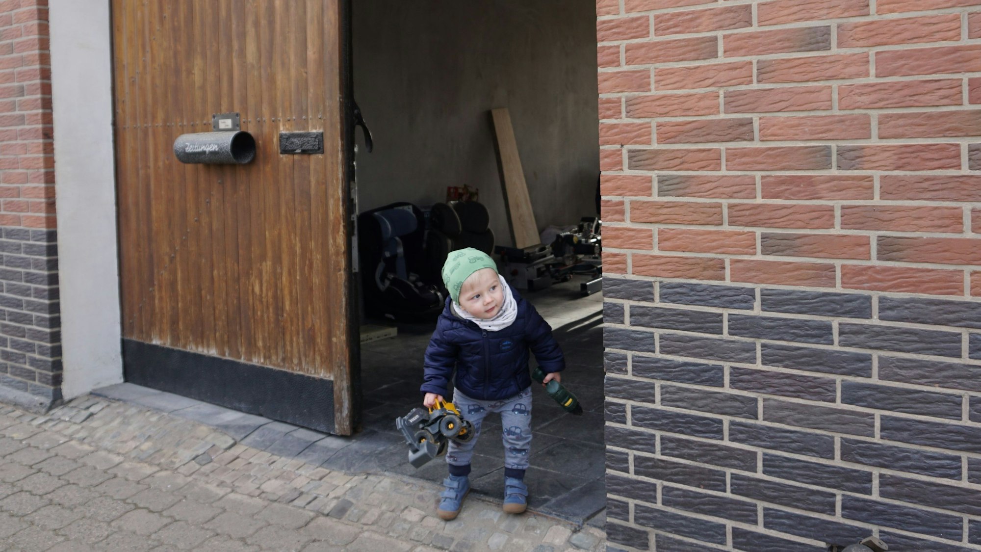 Der kleine Max, einen Spielzeugbagger in der Hand, lugt vorsichtig aus dem Hoftor auf die Straße vor dem Haus.