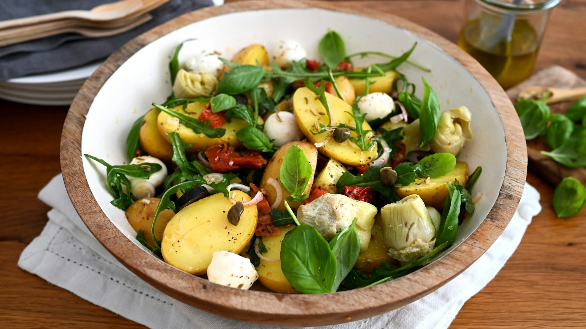 In einer Schüssel sind Kartoffeln, Rucola, Mozzarella, Artischocken, Oliven und getrocknete Tomaten zu einem Salat zubereitet.