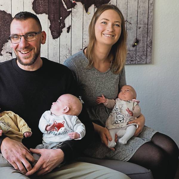Marco Albrecht und Julia Kratzenberg haben ihre drei Babys auf dem Arm und sitzen auf einem Sofa.