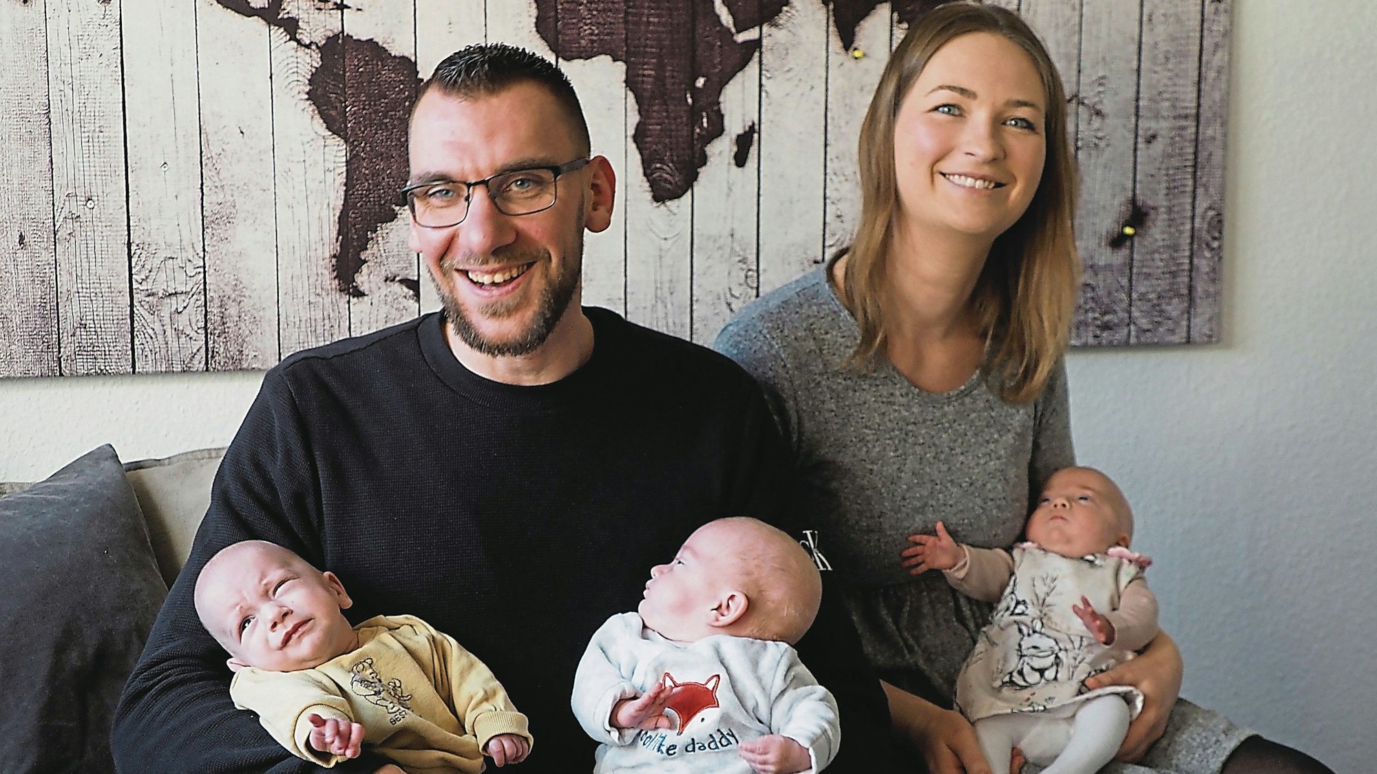 Marco Albrecht und Julia Kratzenberg haben ihre drei Babys auf dem Arm und sitzen auf einem Sofa.