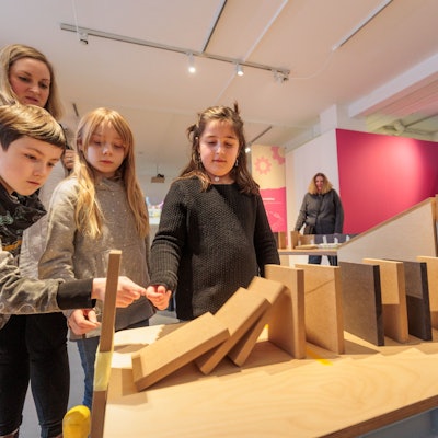 Mehrere Kinder schauen gespannt auf Holzplatten, die als Dominosteine in einer Reihe aufgebaut wurden. Ein Junge stößt den ersten Stein an.