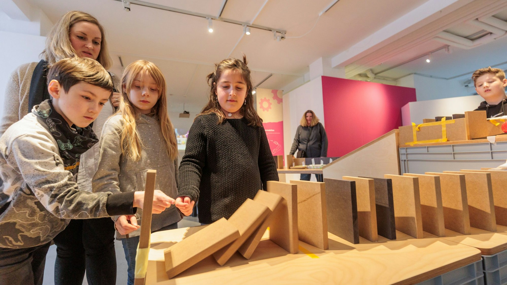 Mehrere Kinder schauen gespannt auf Holzplatten, die als Dominosteine in einer Reihe aufgebaut wurden. Ein Junge stößt den ersten Stein an.