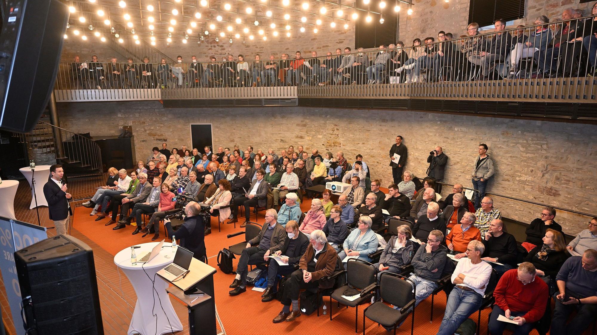 Menschen sitzen im Bensberger Ratssaal und hören einem Sprecher auf der Bühne zu.