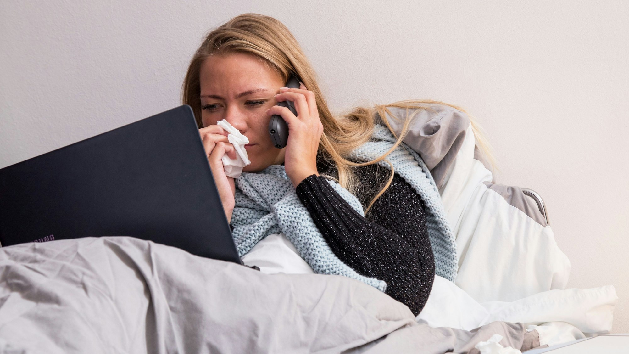 Eine kranke Frau liegt im Bett, telefoniert und hat einen Laptop vor sich.