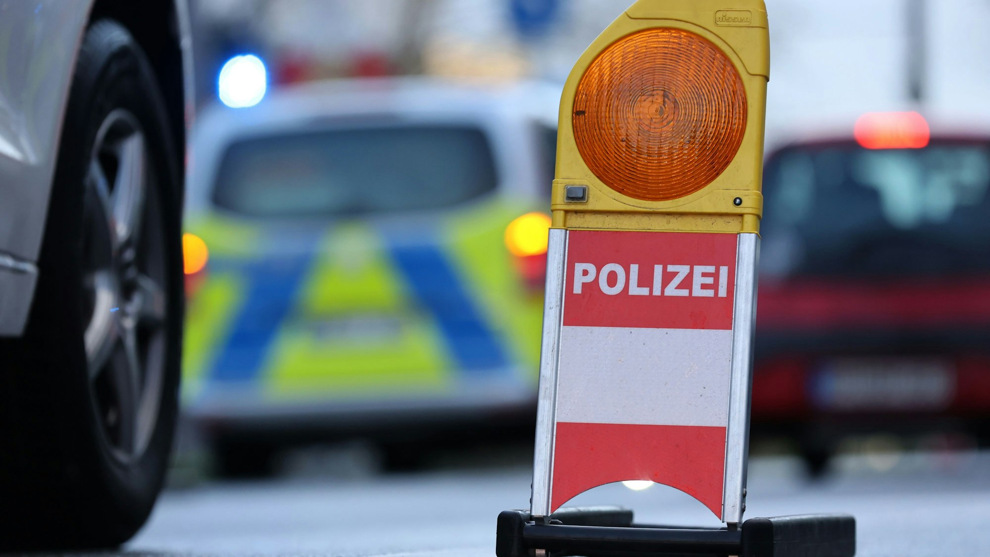 Eine Polizei-Warnbake steht auf einer Straße. Im Hintergrund ist ein Polizeifahrzeug mit Blaulicht zu sehen.