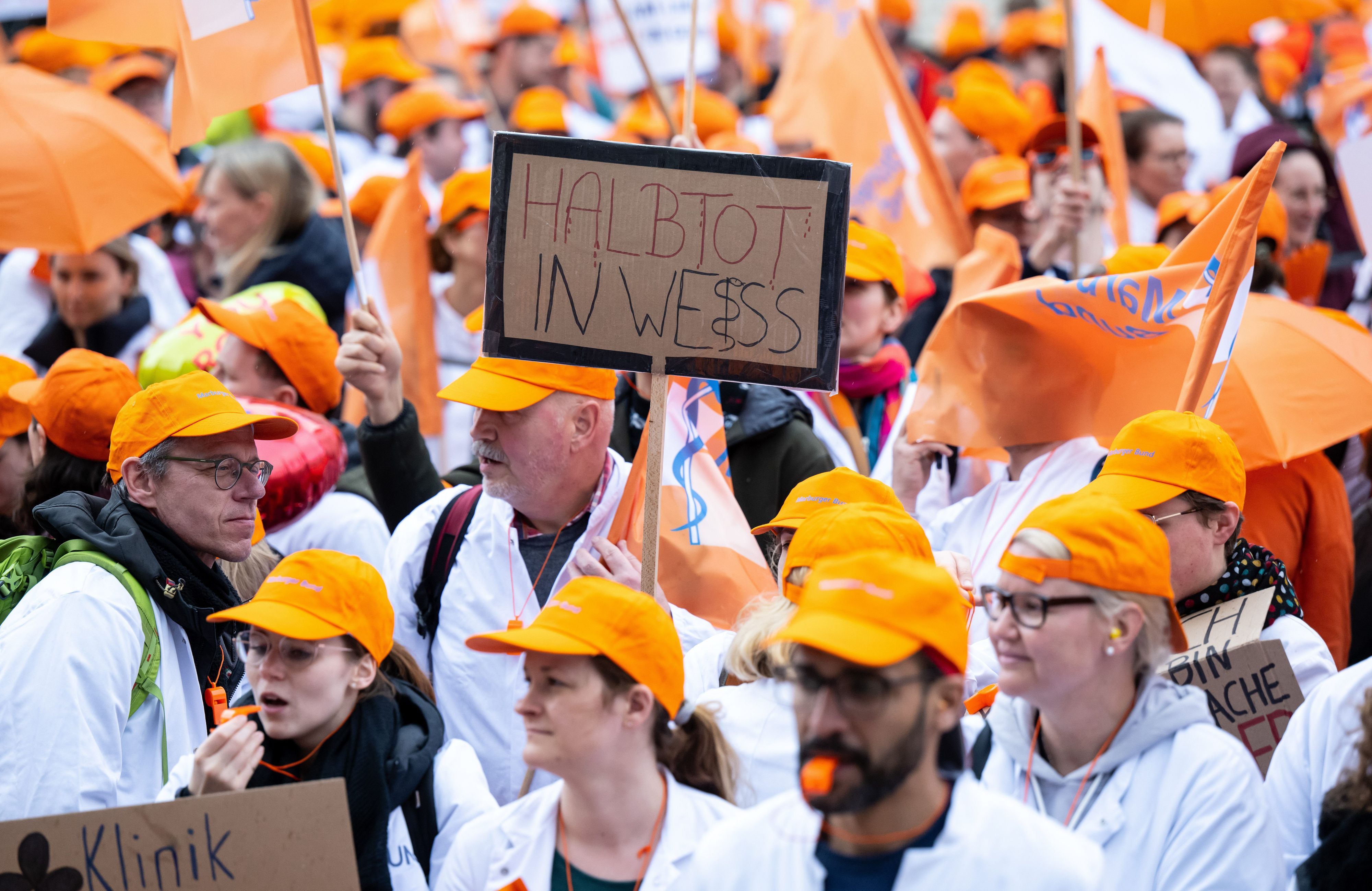 Teilnehmerinnen und Teilnehmer eines Warnstreiks der Gewerkschaft Marburger Bund nehmen an einer Demonstration teil und halten ein Plakat mit der Aufschrift „Halbtot in weiß“ in den Händen.