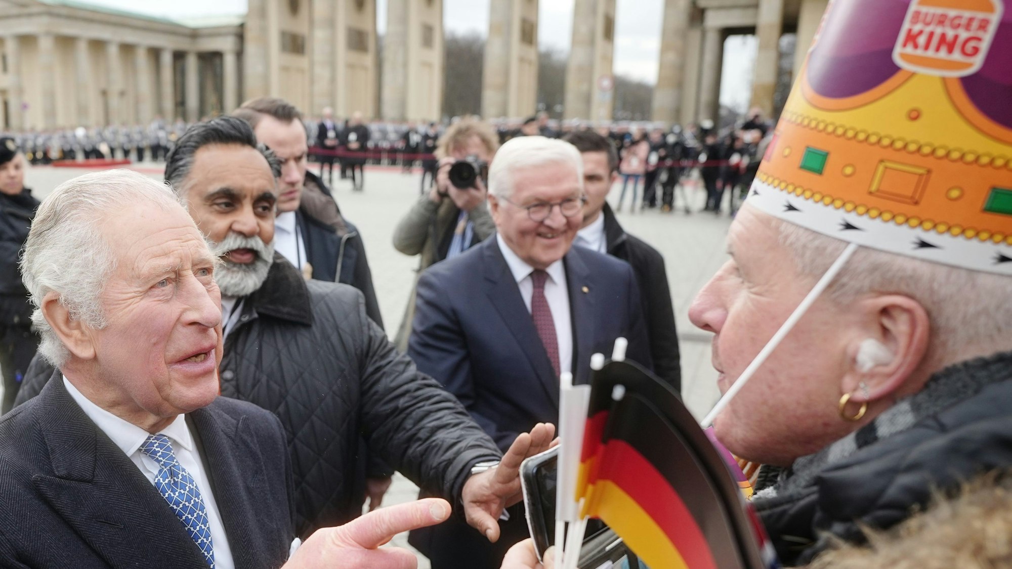 König Charles III. von Großbritannien am Brandenburger Tor mit Bundespräsident Frank-Walter Steinmeier. Ihm gegenüber steht ein Mann mit einer Krone der Fast-Food-Kette „Burger King“.