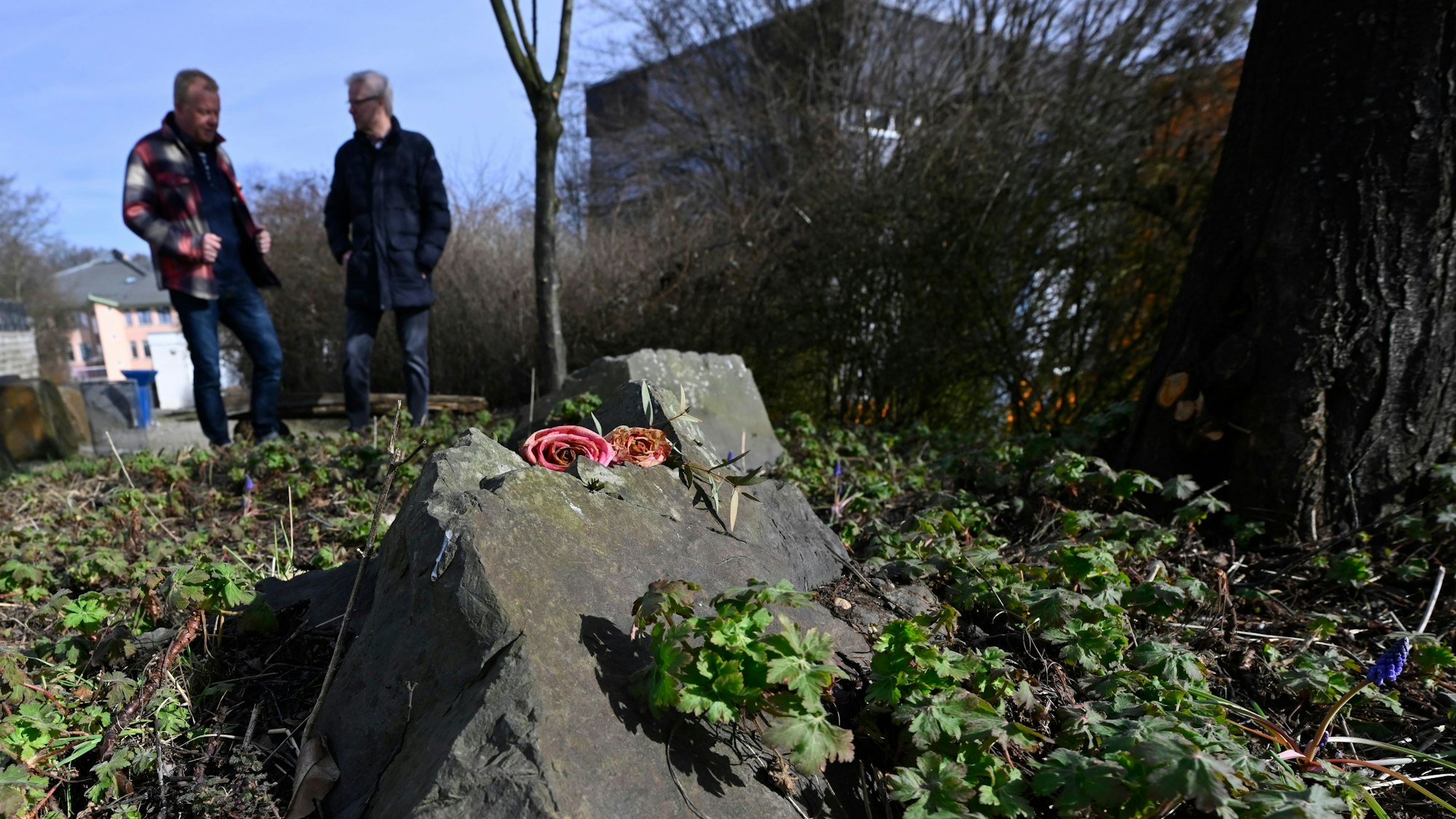 Zwei Männer stehen an einem Gedenkstein, auf dem Rosen liegen.