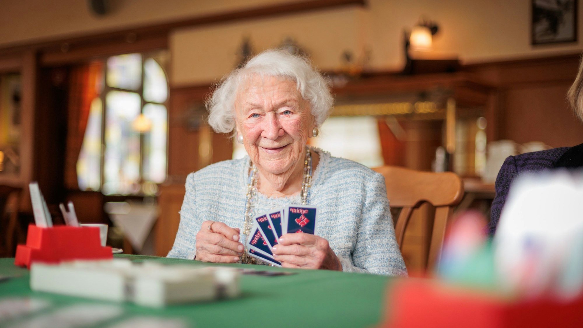 Eine ältere Frau sitzt an einem Tisch und hält Bridge-Karten aufgefächert in der Hand.