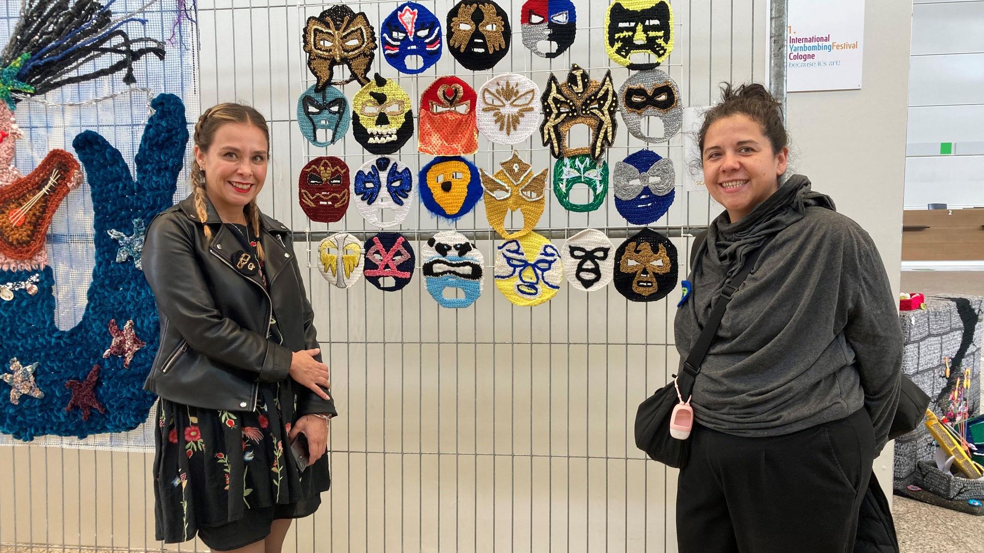 .Zwei Frauen stehen vor einer Wand mit Masken aus Wolle.