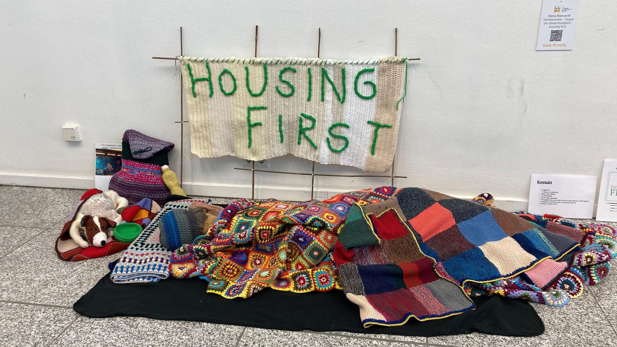 Ein Kunstwerk aus Wolle, das aussieht, wie der Schlafbereich einer obdachlosen Person. Auf einem Banner steht „Housing first“.