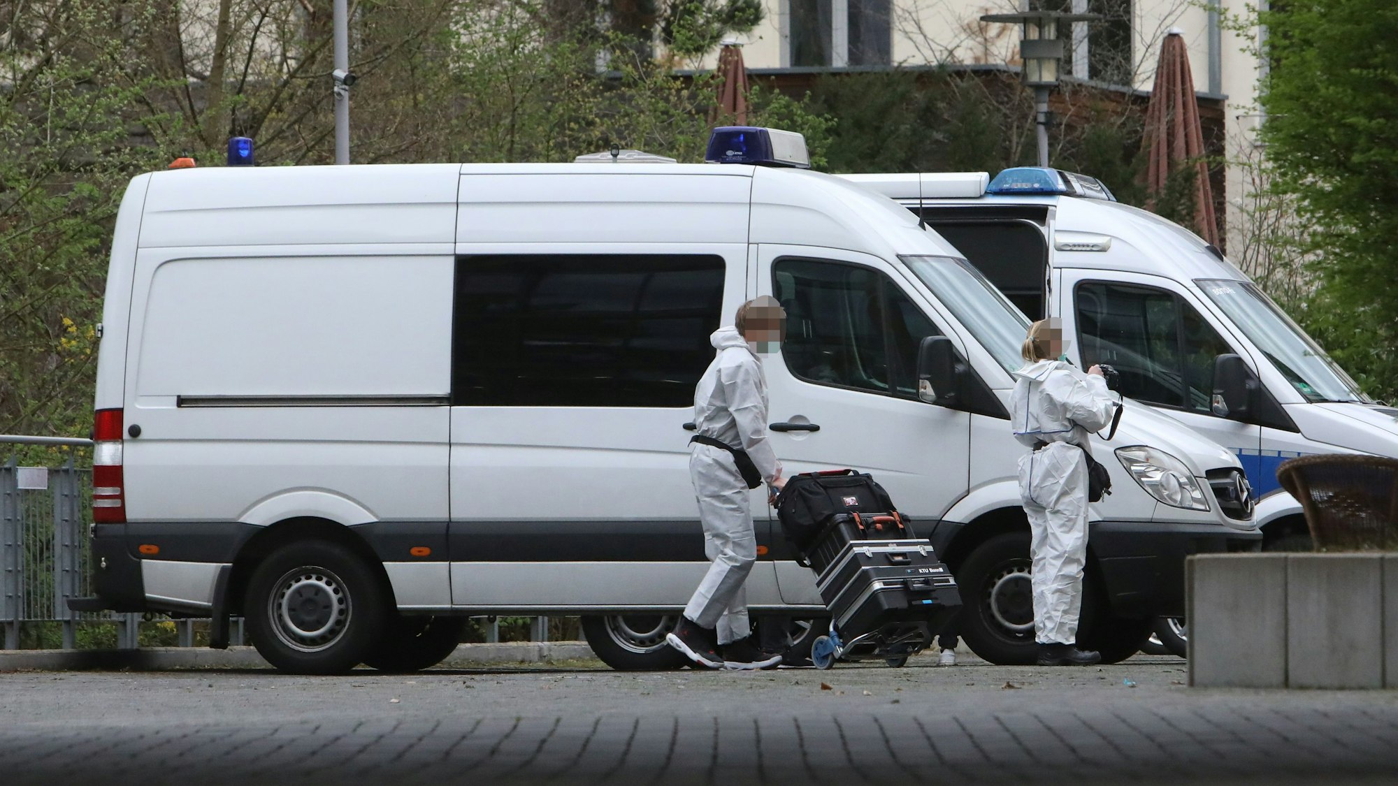 Ein Mann in weißem Schutzzanzug fährt eine mit Koffern beladene Sackkarre von einem Transporter weg, eine Frai im Schutzanzug hält eine Kamera in der Hand.
