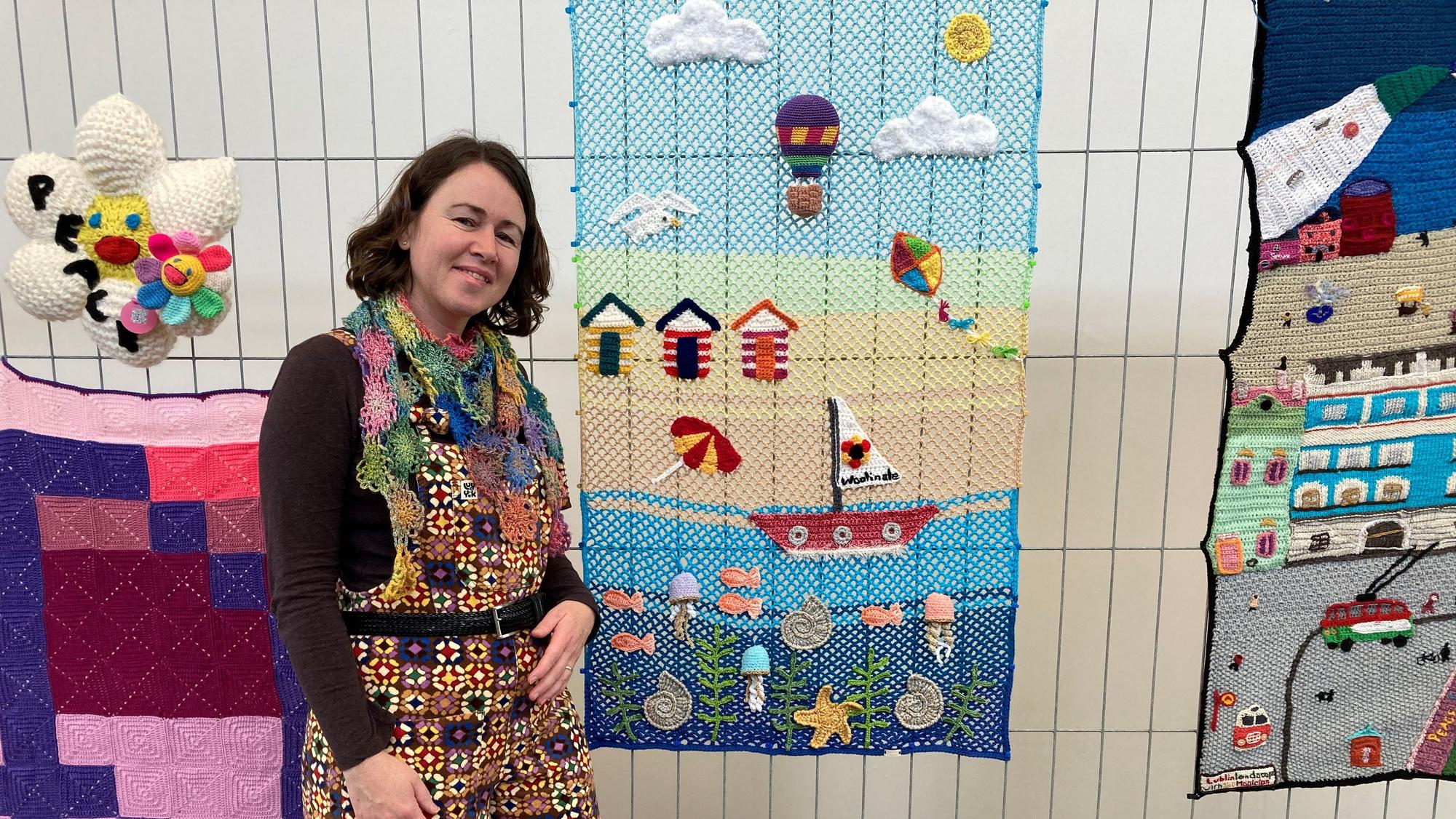 EIne brünette Frau steht aus einem Kunstwerk aus Wolle – Man sieht eine Strandszene mit einem Boot.