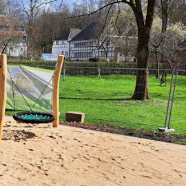 Eine neue Doppelschaukel steht auf einem Sandplatz, noch umgeben von Gitterzäunen. Im Hintergrund sind Fachwerkgebäude des Papiermuseums Alte Dombach zu sehen.